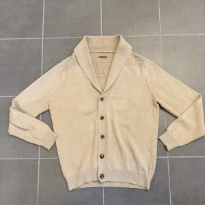 Beige kofta med sjalkrage och knappar från Massimo Dutti - Snygg beige kofta med sjalkrage och fem knappar framtill. Stickad struktur på axlar och krage ger extra detalj. Långa ärmar och ribbade muddar vid ärmslut och nederkant. Perfekt för lager på lager och en clean look.