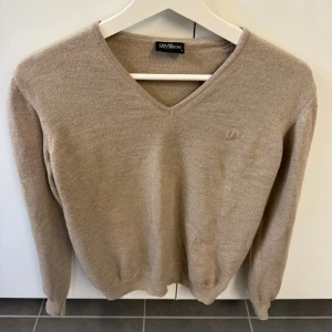 Beige stickad tröja från Oscar Jacobson - Klassisk beige stickad tröja från Oscar Jacobson med V-ringning och diskret broderad logga på bröstet. Tröjan har långa ärmar och ribbade muddar vid ärmslut och nederkant. Perfekt för lager på lager och stilrena outfits.