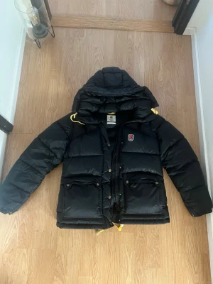 Svart dunjacka från Fjällräven - Svart dunjacka från Fjällräven med huva. Jackan har två stora fickor framtill, dragkedja och tryckknappar. Klassisk Fjällräven-logga. Perfekt för kalla dagar och riktigt varm. Jackan är i fint skick, det ända är ett litet ihopsytt hål på ena armen men det märks inte så tydligt. Endast använd en vinter.