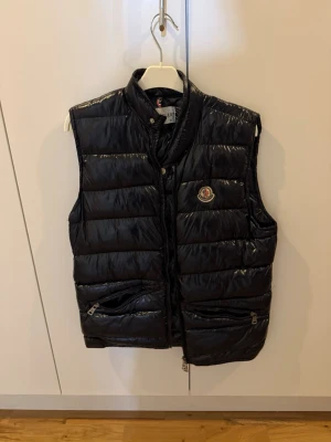 Svart dunväst från Moncler - Snygg svart dunväst från Moncler med glansig finish och ståkrage. Västen har två dragkedjeförsedda fickor framtill och klassisk Moncler-logga på bröstet. Perfekt för lager-på-lager och streetstyle. Materialet är dunfyllning med ett yttertyg i nylon.