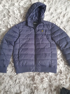Blå pufferjacka från Lyle & Scott - Snygg blå pufferjacka från Lyle & Scott med huva och dragkedja framtill. Jackan har det klassiska gula logotypmärket på bröstet och ribbade muddar vid ärmslut och nederkant. Perfekt för kalla dagar och riktigt skön att bära. Materialet är lätt och quiltat.