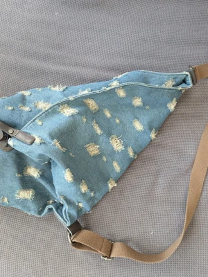 Blå denim crossbody-väska - Säljer en blå crossbody-väska i denim med slitna detaljer över hela väskan. Väskan har en justerbar beige axelrem och dragkedja upptill. Perfekt för dig som gillar en avslappnad och trendig stil.
