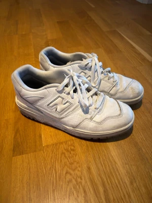 Vita New Balance 550 sneakers - Säljer ett par klassiska vita New Balance 550 sneakers i läder med perforerade detaljer och ikoniska N-loggan på sidan. Skorna har lågt skaft, vita snören och en chunky sula som ger retrovibbar. 