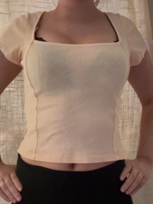 Ljusbeige ribbad topp med kort ärm - Säljer en ljusbeige ribbad topp med fyrkantig urringning och korta ärmar. Toppen har en figurnära passform och markerade sömmar längs sidorna som ger en snygg siluett. Perfekt att styla med både jeans och kjol.