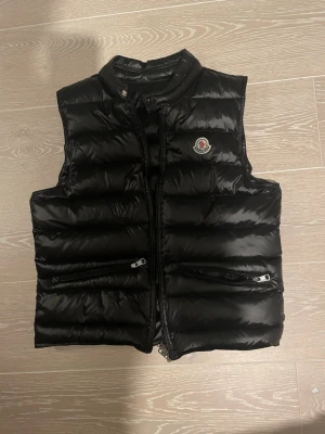 Svart dunväst från Moncler - Snygg svart dunväst från Moncler med glansig finish och klassisk logotyp på bröstet. Västen har ståkrage, dragkedja framtill och två fickor med dragkedja. Perfekt för lager på lager och streetwear-vibe.