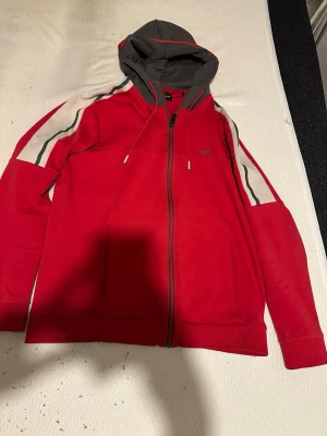 Röd hoodie med dragkedja från BOSS - Snygg röd hoodie från BOSS med dragkedja framtill och kontrasterande mörkgrå huva. Har vita och svarta ränder längs ärmarna och två fickor fram. Tillverkad i mjukt bomullsmaterial och har justerbara snören i huvan. Perfekt för en avslappnad streetwear-look.