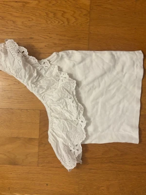 Vit oneshoulder topp med spetsvolang - Säljer en vit oneshoulder topp med bred spetsvolang längs axeln. Toppen är ärmlös på ena sidan och har ett romantiskt uttryck tack vare den broderade spetsen. Perfekt för dig som vill sticka ut lite extra.