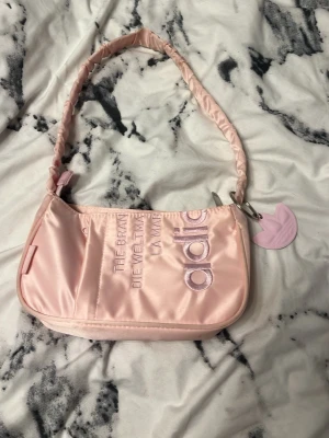 Ljusrosa axelväska från Adidas - Säljer en ljusrosa handväska från Adidas med broderad logga och text på framsidan. Väskan har en vadderad axelrem och en matchande rosa Adidas-berlock. Tillverkad i glansigt syntetmaterial och har en mjuk, sportig look. Pris kan diskuteras, tvivla inte att höra av er eller ställa frågor.💕🩷