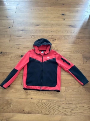 Nike tech fleece - Snygg hoodie från Nike i rött och svart med hel dragkedja framtill och huva. Hoodien har en ficka med dragkedja på ärmen och är tillverkad i ett mjukt, sportigt material. Perfekt för dig som gillar streetwear och sportig stil.