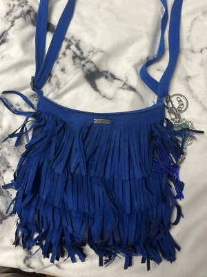 Blå axelväska med fransar Hollister - Säljer en blå axelväska från Hollister med långa fransar på framsidan och en dekorativ nyckelring med hjärta och pärlor. Väskan har justerbar axelrem och är gjord i ett mjukt material som känns som mocka.