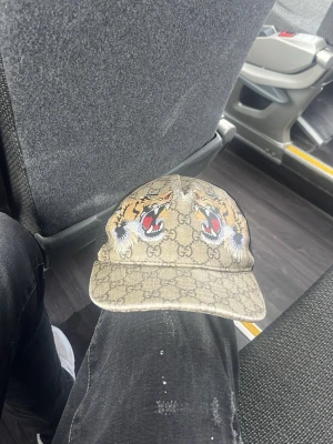 Gucci keps med tigertryck - Säljer en beige Gucci keps med klassiskt GG-mönster och två stora tigerhuvuden framtill. Kepsen har justerbar rem bak med rödgrön rand och är tillverkad i canvasmaterial. Perfekt för dig som vill sticka ut med en lyxig accessoar.