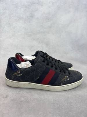 Gucci sneakers med tiger och röd-blå rand - Snygga svarta Gucci sneakers med mörkblå och röda ränder på sidan, diskret GG-mönster i bakgrunden och broderade tigrar. Skorna har vit sula och svarta skosnören. Materialet är canvas med läderdetaljer och klassisk låg profil.