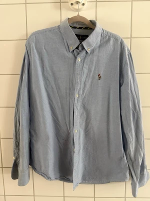 Blå skjorta från Polo Ralph Lauren - Klassisk ljusblå skjorta från Polo Ralph Lauren med broderad logga på bröstet. Skjortan har knappar hela vägen, button-down krage och långa ärmar. Perfekt för en clean och stilren look. I storlek L men passar bättre i M. Skriv om ni har funderingar!