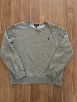 Grå sweatshirt från Polo Ralph Lauren - Klassisk grå sweatshirt från Polo Ralph Lauren med rund halsringning och diskret broderad logga på bröstet. Tröjan har ribbade muddar vid ärmslut och nederkant, perfekt för en avslappnad och stilren look. Storlek XS L (14-16). Bara hör av er vid funderingar! 😃