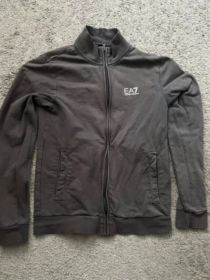 Mörkgrå ziptröja från EA7 Armani - Snygg mörkgrå ziptröja från EA7 Emporio Armani med hög krage och två fickor framtill. Tröjan har en diskret logga på bröstet och är tillverkad i mjukt bomullsmaterial. Perfekt för en avslappnad och stilren look.
