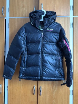 Svart Peak Performance dunjacka - Snygg svart dunjacka från Peak Performance med huva och dragkedja framtill. Jackan har en cool detalj med rosa dragkedja på ärmen och logga på bröstet. Tillverkad i glansigt material och perfekt för kalla vinterdagar. Modellen heter Black Light.