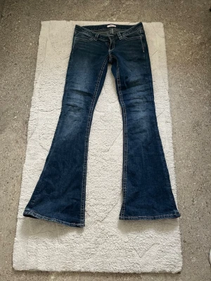 Mörkblå bootcut jeans med broderi - Säljer ett par mörkblå bootcut jeans med snyggt slitna detaljer och broderade bakfickor. Jeansen har låg midja och klassisk femficksmodell. Materialet är denim i bomull med lite stretch för skön passform. Perfekta för dig som gillar retrostil!