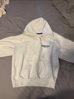 Grå Essentials Fear of God hoodie - Snygg ljusgrå hoodie från Essentials Fear of God med svart logga på bröstet och stor text på ryggen. Klassisk känguruficka framtill och stor huva. Tillverkad i mjukt bomullsmaterial som är perfekt för en avslappnad streetwear-look.