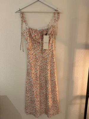 Blommig klänning från Adoore - Supersöt långklänning från Adoore med smala axelband och knyt vid axlarna. Klänningen är vit med små orange/ rosa blommor och gröna blad. Lätt och luftig i materialet.
