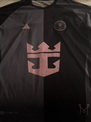 Inter Miami svart fotbollströja Adidas - Snygg svart och mörkgrå fotbollströja från Inter Miami med rosa detaljer och klubbmärke på bröstet. Tröjan har Adidas-logga och stor rosa logotyp på magen. Tillverkad i lätt och ventilerande material, perfekt för träning eller match.