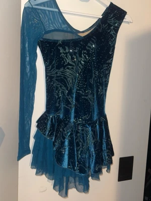 Blå sammetsdräkt med tyllkjol - Snygg blå konståkningsdräkt i sammet med glittrigt mönster och volangkjol. Ena ärmen är lång i mesh och den andra axeln är bar för en cool look. Matchande scrunchie medföljer. Perfekt för träning eller tävling på isen.