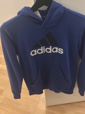 Blå hoodie från Adidas med logga - Snygg blå hoodie från Adidas med stor vit och svart logga på bröstet. Klassisk känguruficka framtill och huva. Tillverkad i mjukt material som passar perfekt för en avslappnad stil. Perfekt för dig som gillar sportig streetwear.litrn fläck vet inte hur ens mrn under luvan därav priset . Annars helt ny. 