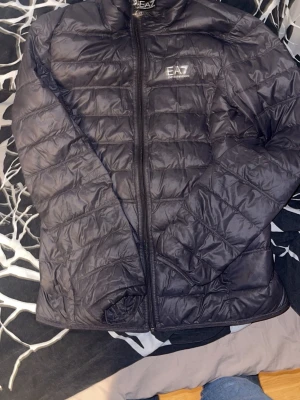 Svart pufferjacka från EA7 Emporio Armani - Säljer en svart pufferjacka från EA7 Emporio Armani med quiltad design och dragkedja framtill. Jackan har hög krage och diskret logga på bröstet. Perfekt för kalla dagar och riktigt snygg till streetwear-stilen.