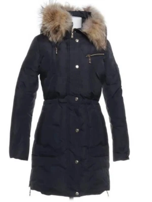 Hollies Svart lång dunjacka med fuskpäls - Snygg svart lång dunjacka med huva och fuskpälsdetalj. Jackan har dragkedja och knappar framtill, samt en bröstficka med dragkedja. Figursydd modell som ger en schysst siluett och håller dig varm under kalla vinterdagar.