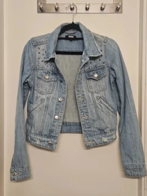 Ljusblå jeansjacka med nitar i nyskick  - Trendig ljusblå jeansjacka från Bik bok med silverfärgade nitar på axlarna och två bröstfickor med lock och knapp. Klassisk krage och knappstängning framtill. Snygga slitningar för en cool look. Perfekt att slänga över en hoodie eller t-shirt. Endast provad så i nyskick.