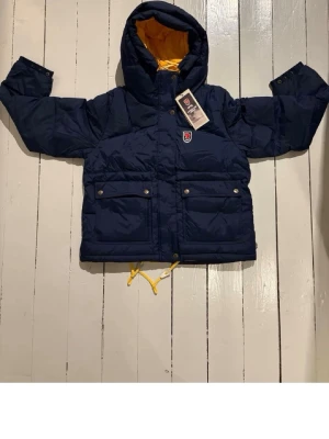 Mörkblå dunjacka från Fjällräven - Oanvänd och helt ny har kvar lappen mörkblå dunjacka från Fjällräven med gul insida på huvan. Säljer då det var fel storlek för mig, hör av er vid fler frågor eller diskutera pris🥰