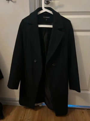 Svart kappa från Kappahl - Stilren svart kappa från Kappahl med klassisk krage och dubbelknäppning framtill. Kappan har raka ärmar och en rak passform, perfekt för en trendig och tidlös look. Fodrad insida för extra komfort.