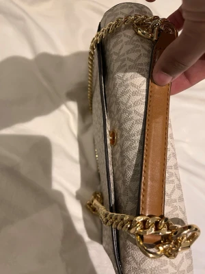 Michael Kors beige handväska - Snygg beige handväska från Michael Kors med klassiskt logomönster och detaljer i brunt läder. Väskan har guldfärgad kedja som axelband och flera fack, inklusive ett med dragkedja. Perfekt för dig som gillar stilrena accessoarer.