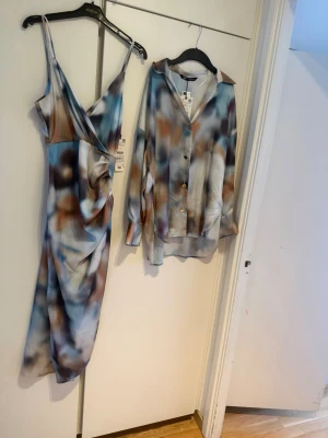 Nytt! Zara set klänning + skjorta i satin mönstrad strl Xs/s - Säljer en snygg omlottklänning från NA-KD med abstrakt mönster i blått, brunt, vitt och beige. Klänningen har smala axelband, draperad midja och v-ringning. Perfekt för dig som gillar trendiga och unika plagg. Strl xs på klänningen och S på skjortan