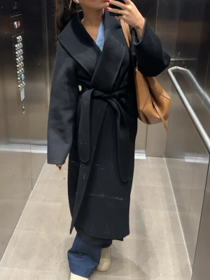 Svart lång kappa  - Stilren svart lång kappa med bred krage och knytskärp i midjan. Kappan har en rak passform och vida ärmar, vilket ger en avslappnad men ändå elegant look. Perfekt för kalla dagar och enkel att matcha med olika outfits.