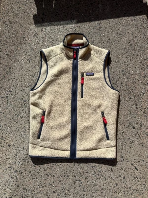Beige fleeceväst från Patagonia - Säljer en beige fleeceväst från Patagonia med marinblå detaljer och röda dragkedjor. Västen har hög krage, tre fickor med dragkedja och är tillverkad i mjukt teddyfleece-material. Perfekt för lager-på-lager och outdoor-vibe.