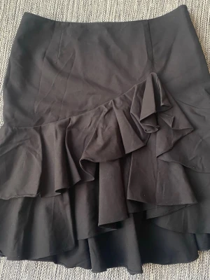Svart volangkjol med asymmetrisk detalj Ralph Lauren - Säljer en svart kortkjol med dubbla volanger och asymmetrisk design. Kjolen har en figurnära passform och är tillverkad i ett mjukt bomullsliknande material. Perfekt för dig som gillar trendiga detaljer och vill sticka ut lite extra. Svart kjol med volanger i amerikansk strl 10 (40) från Ralph Lauren, modell Pauldina Oesant skirt  Sparsamt använd och i fint begagnat skick   Mått Midjan: ca 40 cm Total längd: ca 54 cm  59% bomull, 37% viskos och 4% elastan  Kommer från ett djur och rökfritt hem 