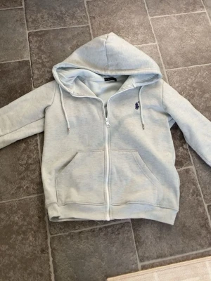 Ljusgrå zip hoodie från Polo Ralph Lauren - Snygg ljusgrå hoodie med dragkedja från Polo Ralph Lauren. Klassisk design med huva och dragsnören, två stora fickor framtill och den ikoniska lilla loggan broderad på bröstet. Perfekt för en avslappnad och stilren look.