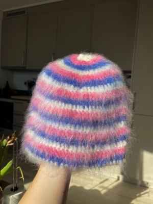 Randig fluffig mohairmössa - Säljer en virkad mössa i mohair med fluffig känsla. Mössan har breda ränder i rosa, blått och vitt som ger en färgglad och lekfull look. Perfekt för dig som vill ha en unik accessoar till vintern. 
