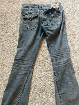 Blå bootcut jeans från True Religion - Säljer ett par ljusblå bootcut jeans från True Religion med ikoniska fickor och vita kontrastsömmar. Jeansen har två bakfickor med lock och knapp, samt klassisk True Religion-lapp bak i midjan. Tillverkade i mjuk denim med snygg tvätt. Köpte för ca 1400kr och är knappt använda. Köpare står för frakt 