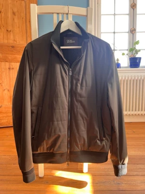 Svart bomberjacka från Oscar Jacobson - Snygg svart bomberjacka från Oscar Jacobson med diskret quiltad design, ribbade muddar och krage. Jackan har dragkedja framtill och två sidofickor med dragkedja. Materialet är slätt och ger en stilren look som passar till många outfits.