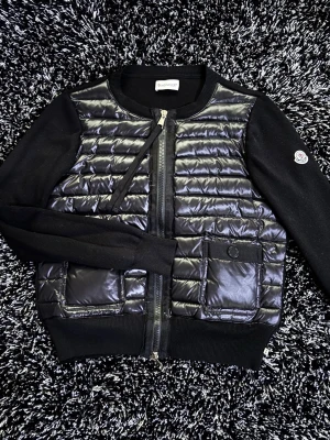 Moncler cardigan - Snygg svart cardiganjacka från Moncler med quiltad, glansig front och två fickor. Ärmarna är i mjukt stickat material och har Moncler-logga på ena ärmen. Dragkedja framtill och ribbade muddar för en clean look. Jackan används både för man och kvinna