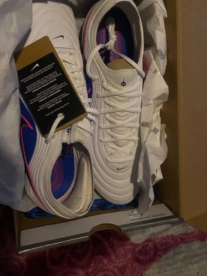 Nike vita och blå fotbollsskor - Säljer ett par Nike fotbollsskor i vitt och blått med stora svarta och rosa swoosh-loggor på sidorna. Skorna har snörning, modern design och är tillverkade i syntetmaterial för bästa grepp och känsla på planen. Perfekta för dig som vill sticka ut på matchen.