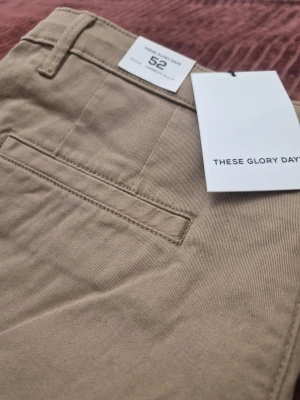 THESE GLORY DAYS T Aimon Tapered beiga chinos, storlek 52 - Snygga beige chinos från These Glory Days i klassisk rak modell. Byxorna har bälteshällor, två sidofickor och två bakfickor med minimalistisk design. Tillverkade i ett mjukt bomullstyg som ger en clean och stilren look.