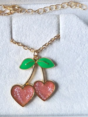 Halsband med körsbärshänge i guld - Sött halsband med ett körsbärshänge i form av två rosa hjärtan med glittereffekt och gröna blad. Kedjan är guldfärgad och hänget har en lekfull och trendig design. Perfekt accessoar för att addera lite färg och personlighet till din outfit.