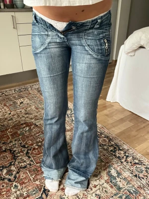Blå low waist bootcut jeans - Säljer ett par blå low waist jeans med bootcut-modell och markerade sömmar. Jeansen har en unik sned knäppning, stora framfickor med dragkedjedetalj och lätt slitna partier för en cool vintagekänsla. Perfekta för dig som gillar Y2K-stil.