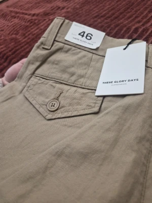 These Glory Days T Fairfox Cargo beige byxor, storlek 46 - Snygga beige cargobyxor från These Glory Days med raka ben och klassiska cargofickor på sidorna. Byxorna har bälteshällor, knappstängning och bakficka med lock och knapp. Tillverkade i ett mjukt bomullstyg som känns skönt mot huden.