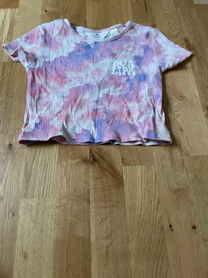 Tie-dye croppad t-shirt från H&M - Säljer en ribbad croppad t-shirt från H&M med tie-dye mönster i rosa, lila och vitt. T-shirten har korta ärmar och ett tryck med texten 'Good Vibes' på bröstet. Perfekt för dig som gillar färgglada och trendiga plagg.