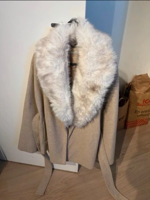 Beige kappa med fuskpäls och bälte - Säljer en beige kappa med stor, fluffig fuskpäls runt kragen och knytskärp i midjan. Kappan har en avslappnad passform och är perfekt för kyliga dagar. Enkel men elegant design som ger en lyxig känsla. Den är köpt på Gina Tricot med nypris 1099 kr och kommer sälja den för runt 800 kr då den knappt är använd 💞Men det går att diskutera priset alltid!