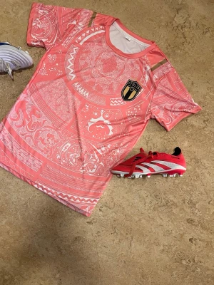 Rosa fotbollströja Italia FC - Säljer en rosa fotbollströja från Italia FC med unikt vitt mönster över hela tröjan och klubbmärke på bröstet. Tröjan har rund hals och korta ärmar, perfekt för träning eller match. Materialet är lätt och andas, vilket gör den skön att spela i.