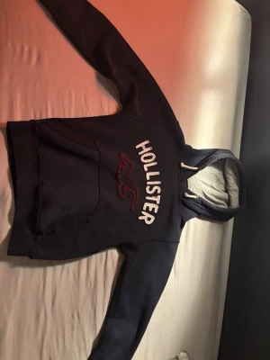 Mörkblå Hollister hoodie - Mörkblå hoodie från Hollister med stor vit broderad logga och röd fågel på bröstet. Den är använd så det finns ett allmänt slitage men inga hål eller liknande. Det står storlek M men passar som XS.